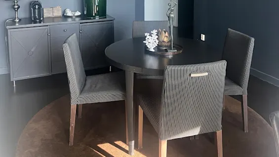 A round black table with striped chairs sits on a brown rug under a chandelier; a cabinet with decor including a plant and lamp is against a dark wall with framed art.