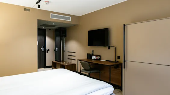 En pænt redt seng står i forgrunden af et moderne hotelværelse med beige vægge. Et vægmonteret TV vender mod sengen over et skrivebord i træ med stol og lampe.