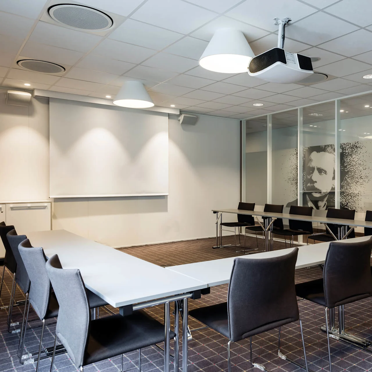 Konferanserom med U-formet bordoppsett og svarte stoler. En projektor henger fra taket, og et veggstort portrett av en mann er synlig i glasspanelene.