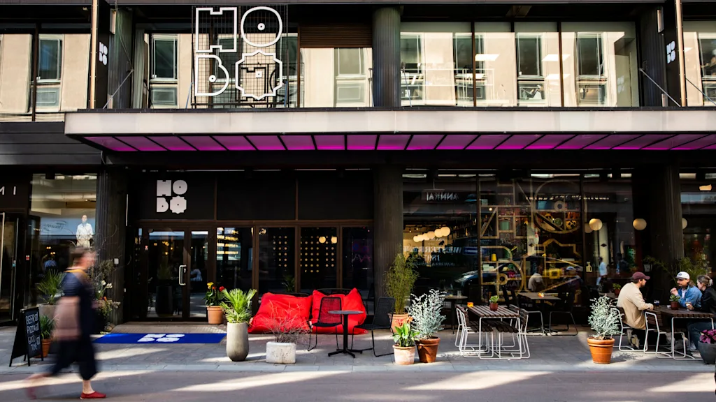 A modern building facade displaying "HO BO" signage. Red seating and potted plants adorn the outdoor area where people are gathered. A person walks past the scene.
