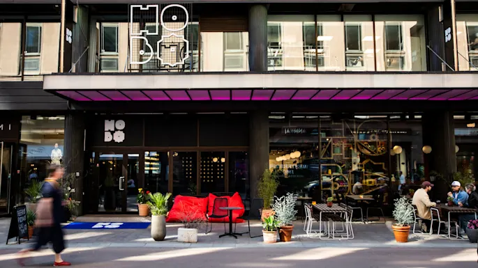 En moderne bygningsfasade med skiltet "HO BO". Røde sitteplasser og potteplanter pynter uteområdet hvor folk samles. En person går forbi.