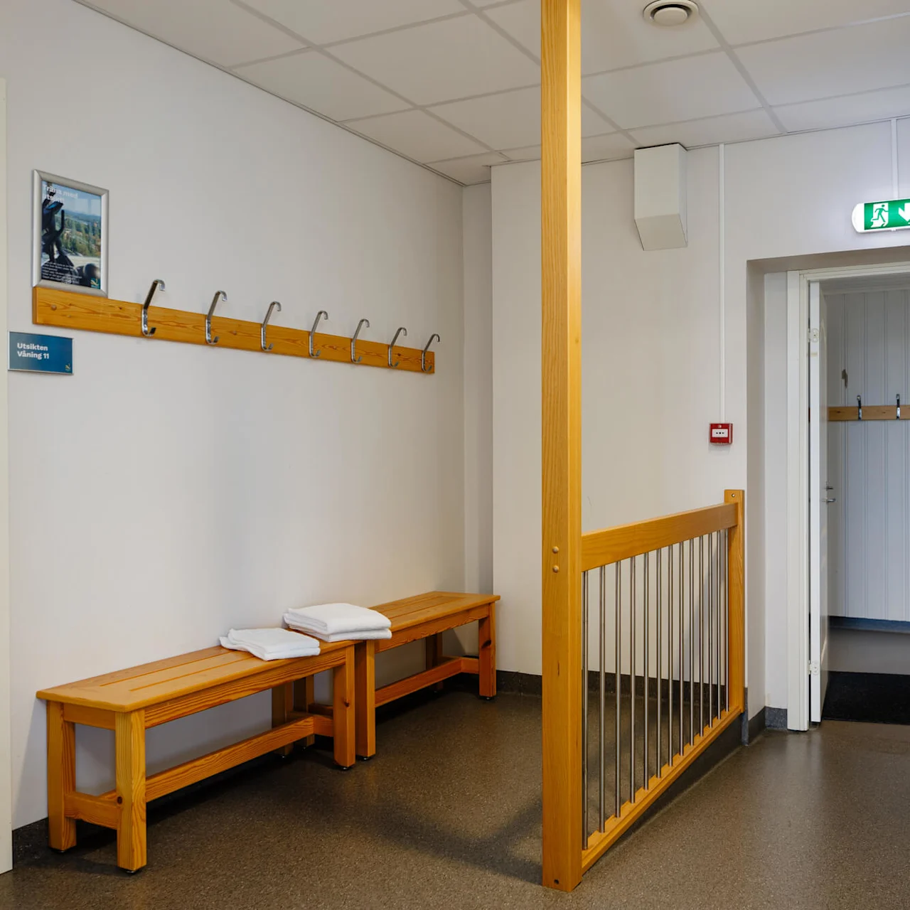 Bänkar med vikta handdukar står mot en vägg med klädkrokar och en bild. En dörr leder till ett annat rum. Skyltar lyder "Utsikten Vikingnäs" och en grön utgång.