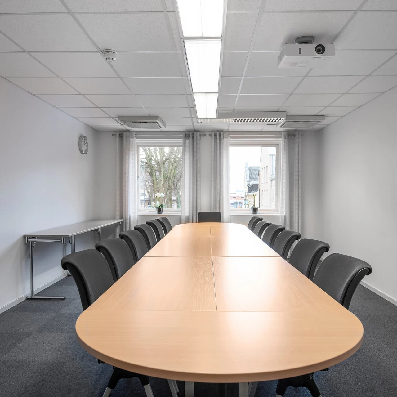 Ett långt konferensbord med svarta stolar runt det, placerat i ett ljust rum med två stora fönster, en klocka på väggen och en projektor i taket.