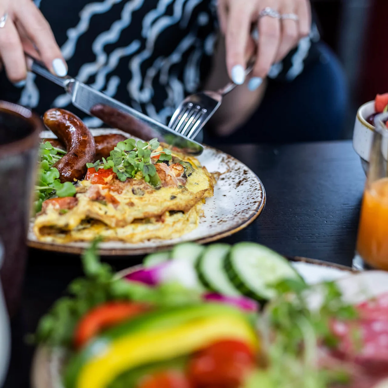 En person skär en omelett med korv och grönt på en tallrik. Runtomkring finns glas med apelsinjuice och färgglada grönsaker på ett mörkt bord.