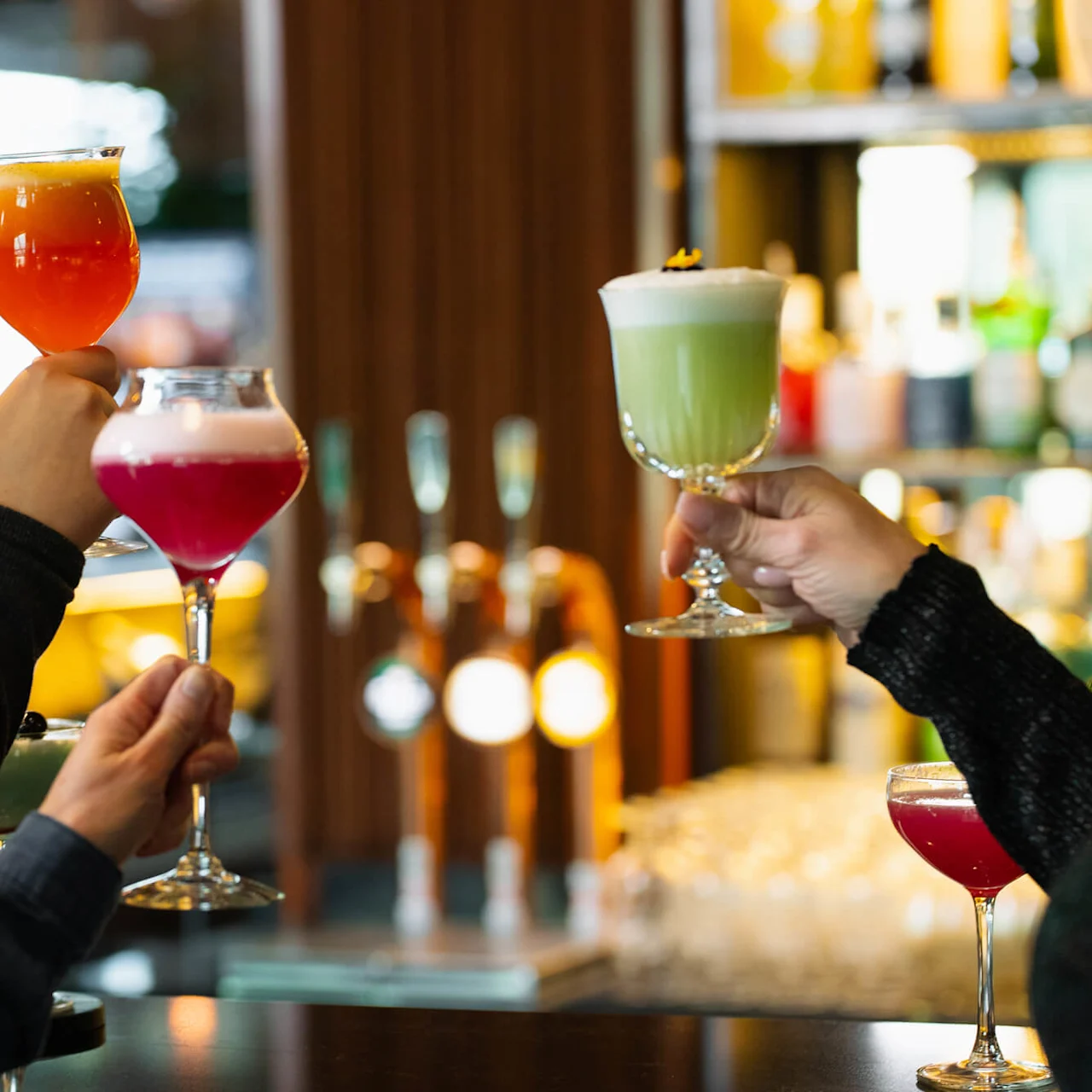 Tre personer håller färgglada cocktails i en bar, med suddiga flaskor och kranar i bakgrunden, vilket skapar en livlig atmosfär.