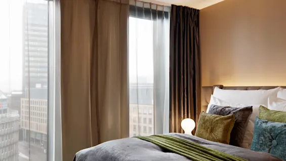 A bed with colorful pillows and a green throw lies by large windows under sheer curtains in a modern hotel room. Outside, tall buildings rise in the cityscape.