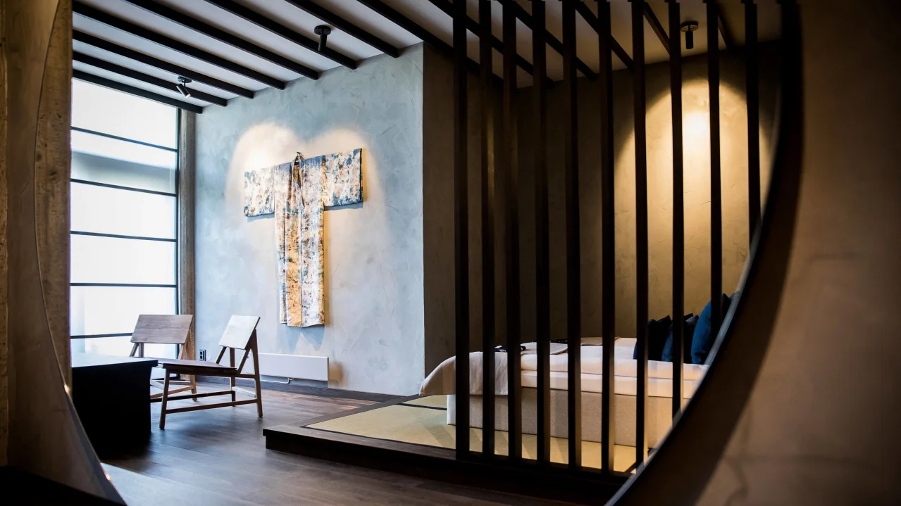 A modern bedroom viewed through an oval mirror, featuring a kimono on the wall and wooden slats separating the sleeping area.