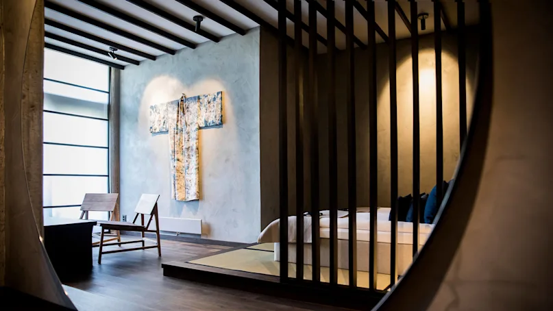 A modern bedroom viewed through an oval mirror, featuring a kimono on the wall and wooden slats separating the sleeping area.