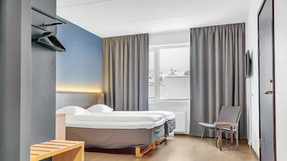A tidy hotel room with twin beds, a chair, mounted TV, and open curtains revealing daylight.