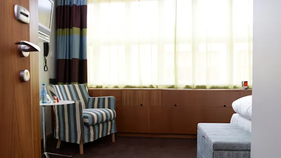 A striped armchair sits beside a small round table with water and chocolates, in a softly lit room with brown carpeting, wooden storage, and sheer curtains filtering sunlight.