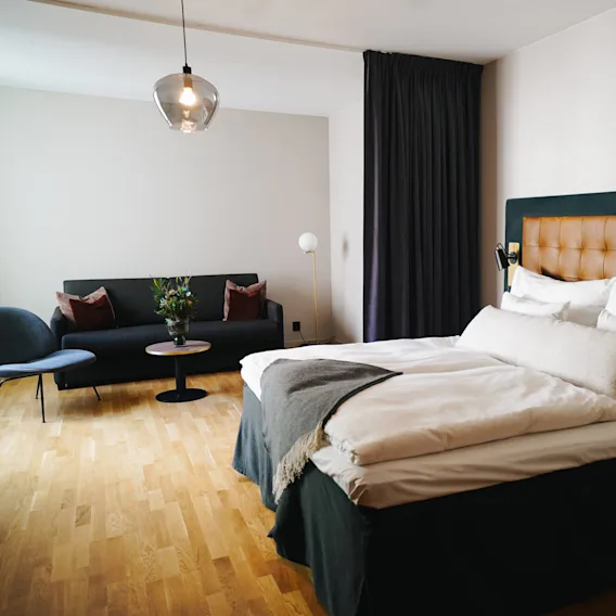 En prydligt bäddad säng med vita lakan och en vadderad sänggavel står i ett rymligt rum. I närheten står en soffa med kuddar mot ett soffbord med en växt, under en hängande lampa.