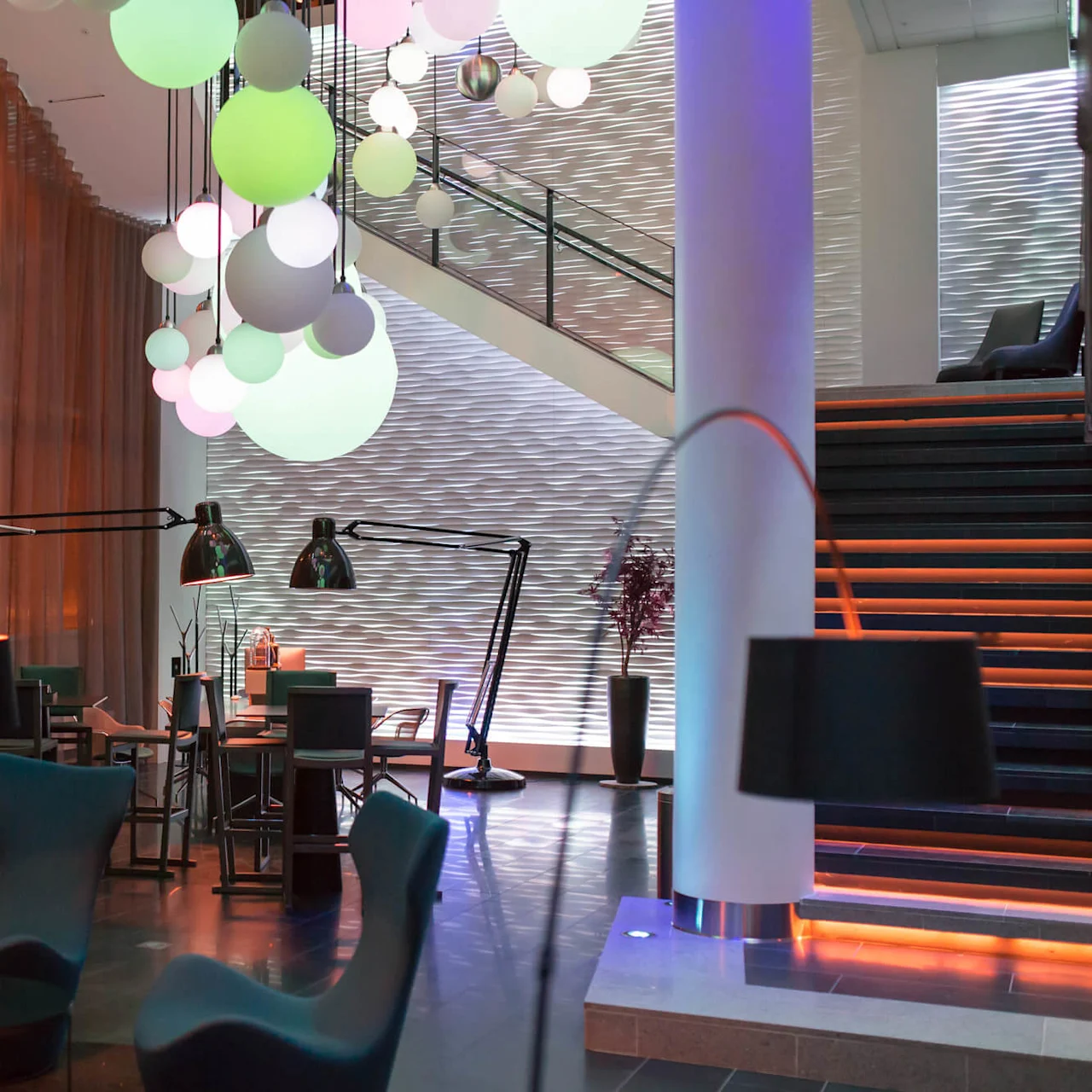 Elegant lounge med moderna stolar och bord, folk samtalar, färgglada klot hänger ovan, upplysta trappor bakom, och varm omgivande belysning genom tunna gardiner.