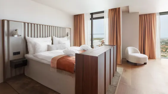 En snyggt bäddad säng med vita lakan och en orange pläd står vid ett fönster i ett modernt hotellrum. Bruna gardiner ramar in en utsikt över ett kuststadsskap.