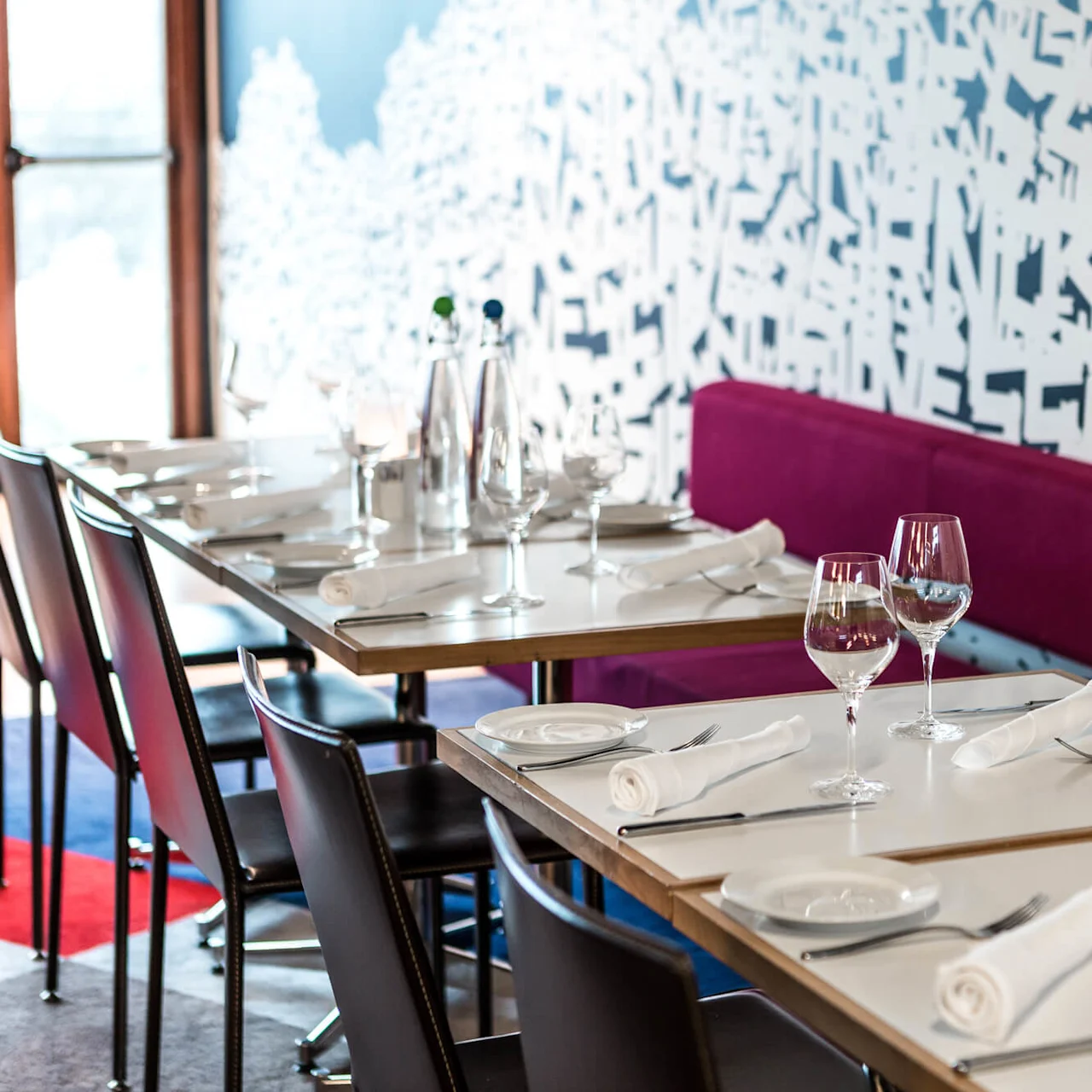 Tables set with wine glasses and napkins, positioned beside a purple upholstered bench in a modern restaurant. Abstract white and blue graffiti-style wall decor adds artistic flair. No visible text.