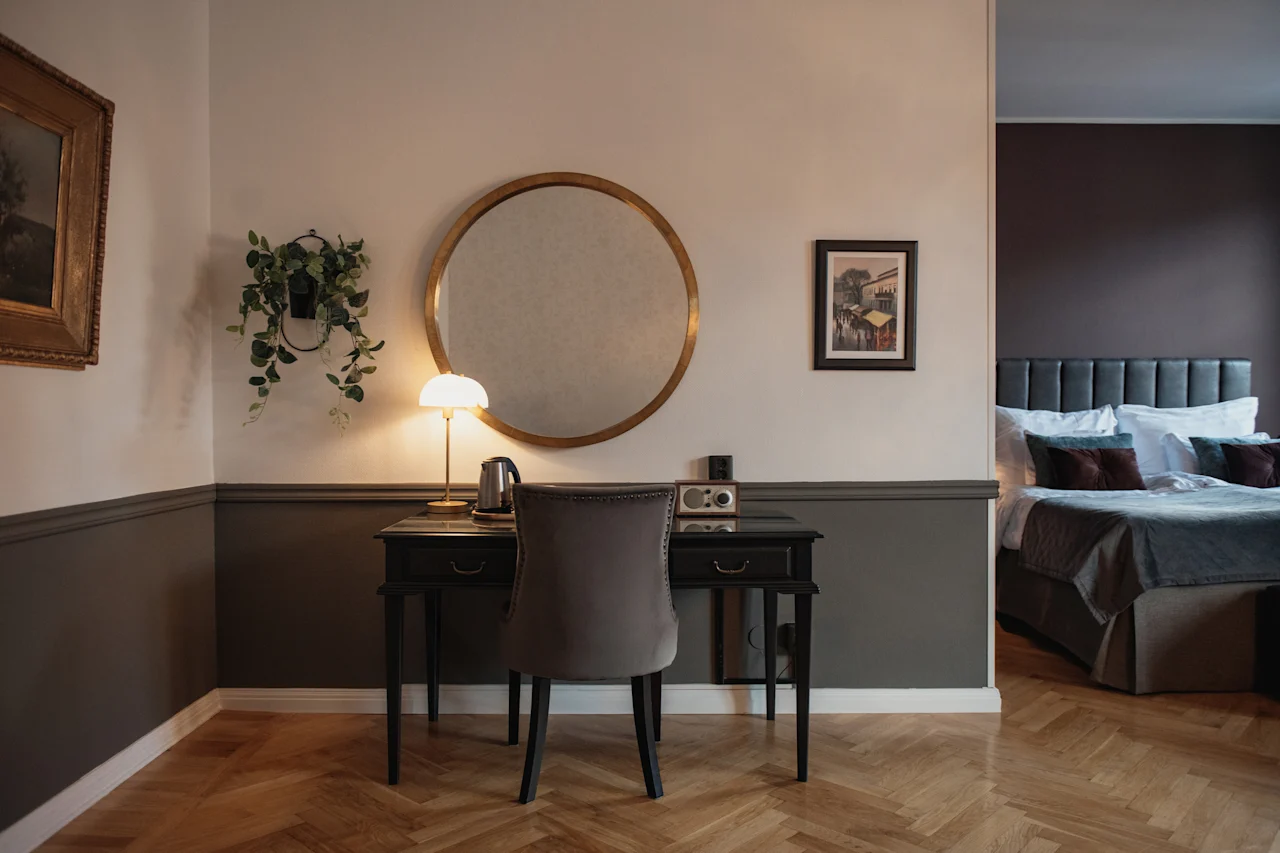 Ett hotellrum med en dedikerad arbetsplats med skrivbord, stol och en stor rund spegel. En säng syns i bakgrunden.