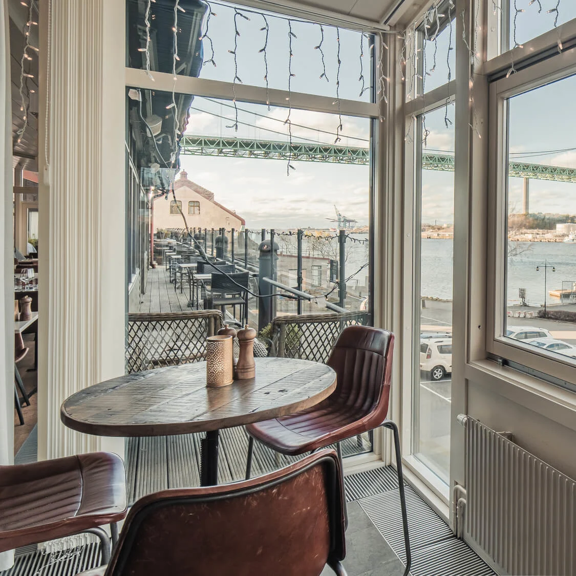 A round wooden table with two chairs overlooks a scenic waterfront view through large windows, with a bridge and buildings visible outside. Several cars are parked by the waterfront.