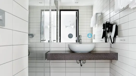 A white sink sits on a dark countertop in a modern bathroom, with hairdryers mounted on a tiled wall. Two stickers read "BE EASY ON THE PLANET" and "GREEN MAKES ME SEE."