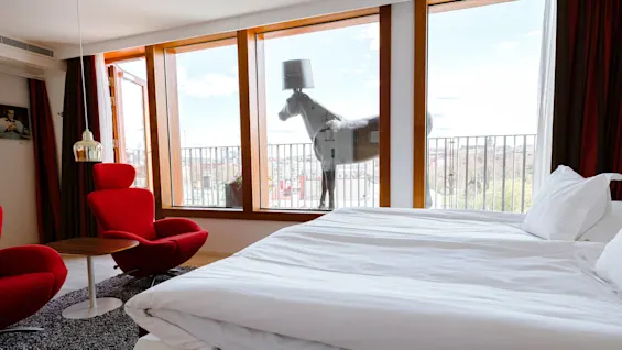 A large bed with white linens is in a modern room. Two red chairs and a small table sit by wide windows overlooking a balcony with a horse statue.