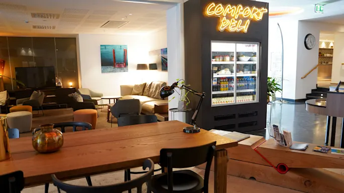 Et moderne venterom med komfortable sitteplasser, et neonskilt som leser "COMFORT DELI," og et velassortert kjøleskap, som skaper en koselig atmosfære.