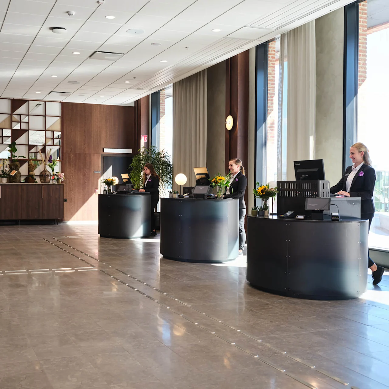 Tre receptionister står bag buede skriveborde og arbejder ved computere i en lyst oplyst hotellobby. En skænk med planter og geometrisk vægdesign pynter i baggrunden og forstærker den moderne stemning.