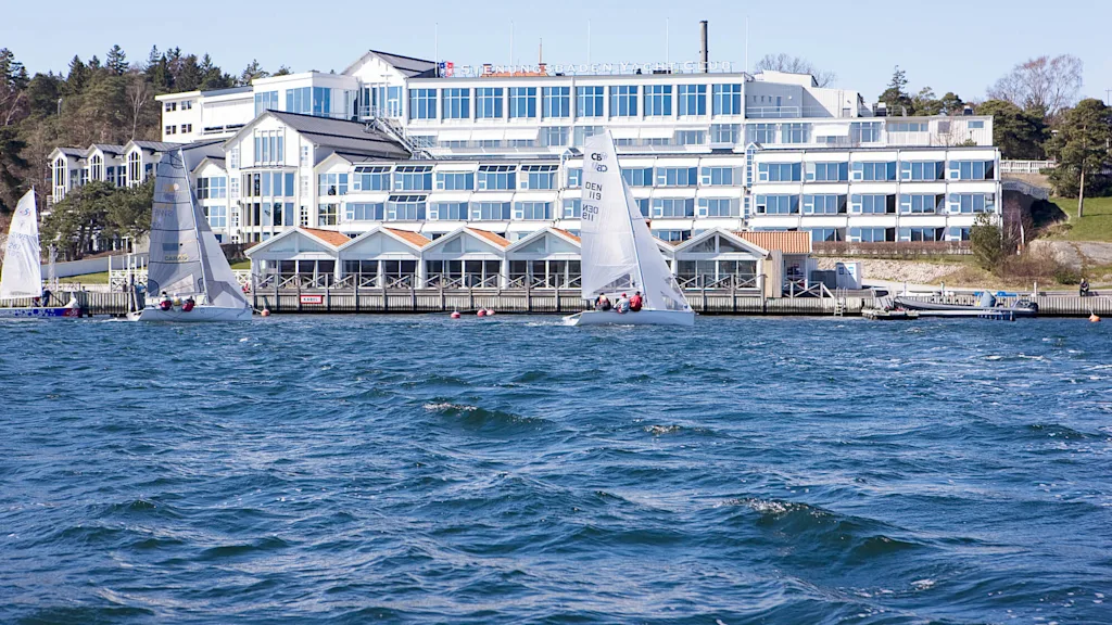 Segelbåtar glider över en solig, blåsig hamn framför en vit, flerplansbyggnad märkt "Svendborg Sunds Sejlklub." Omkringliggande träd skapar en lugn miljö vid vattnet.