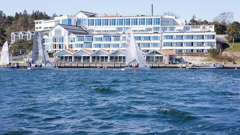 Sailboats glide across a sunny, windy harbor in front of a white, multi-story building labeled "Svendborg Sunds Sejlklub." Surrounding trees create a serene waterfront setting.