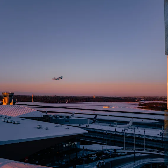 Clarion Hotel Helsinki Airport. Lentokone nousee lumiselta lentokentän kiitotieltä auringonlaskun aikaan, moderni rakennus etualalla.