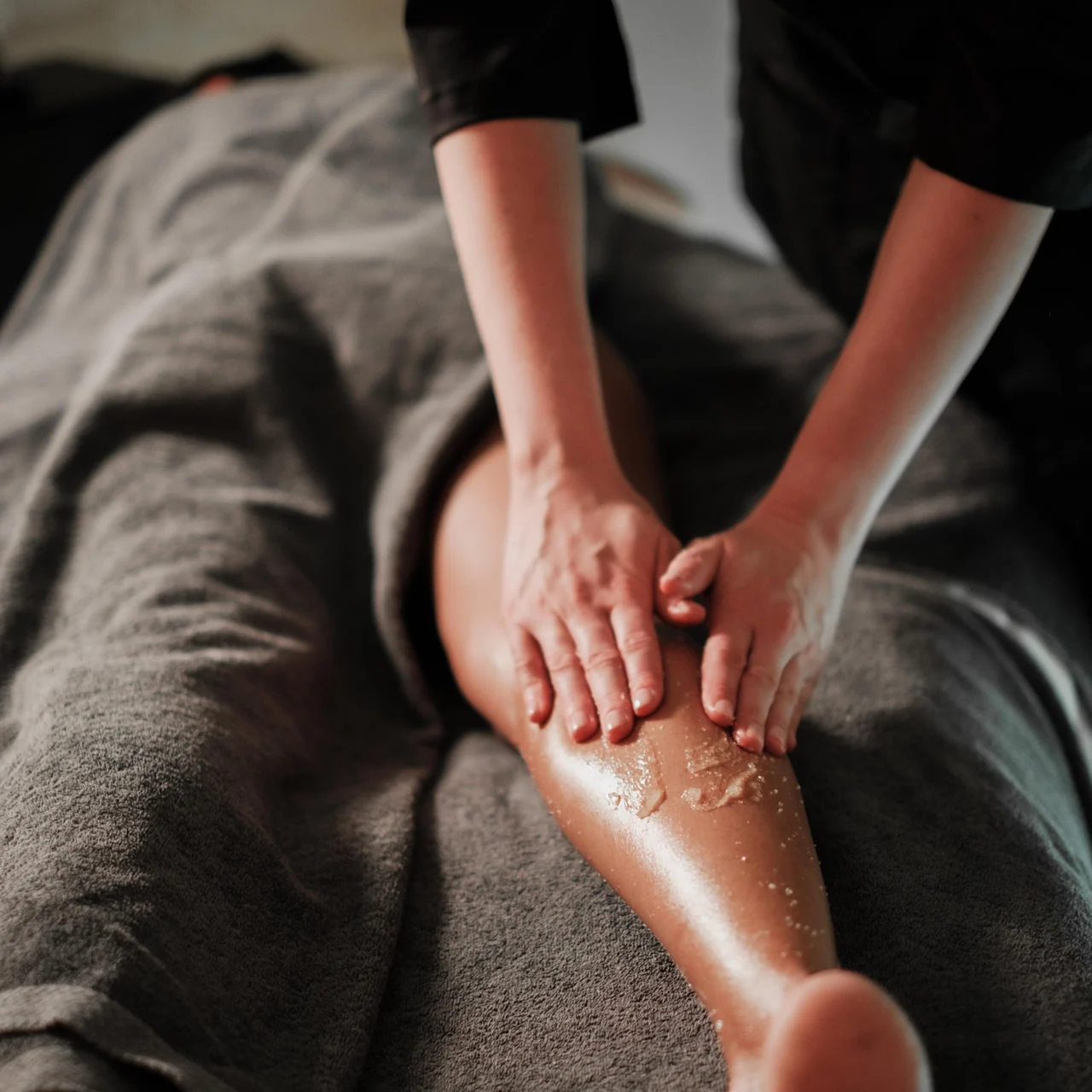 En massageterapeuts händer gnider in ett exfolierande skrubb på en klients ben, som ligger täckt med en grå handduk i en svagt upplyst miljö.