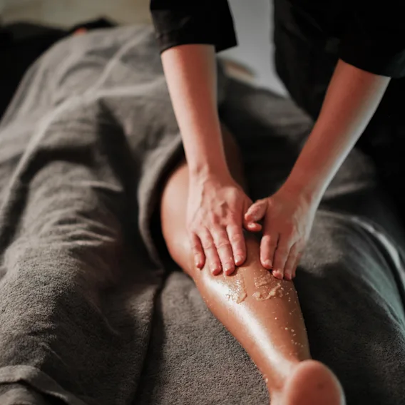 En massageterapeuts händer gnider in ett exfolierande skrubb på en klients ben, som ligger täckt med en grå handduk i en svagt upplyst miljö.