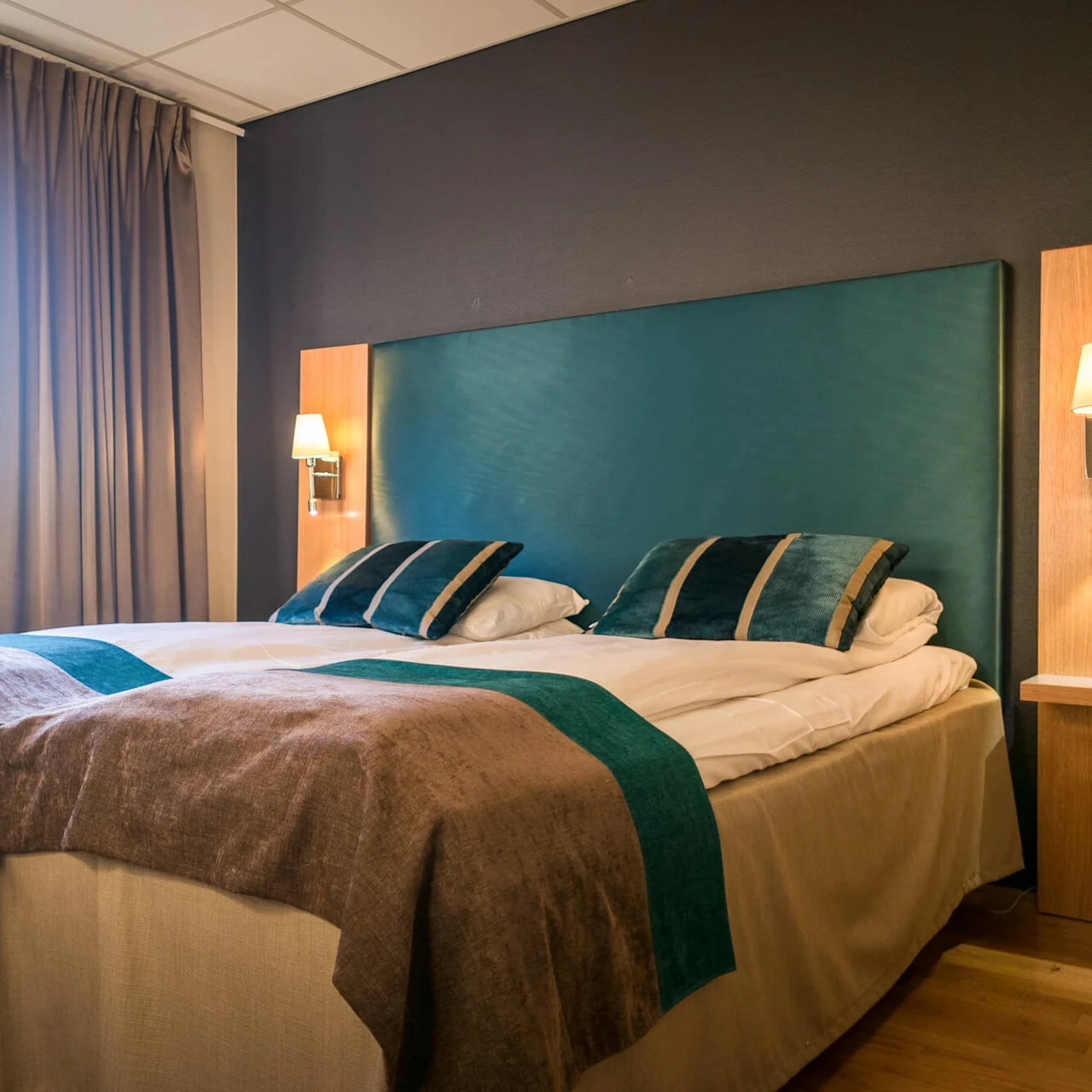 A neatly made hotel bed with teal and brown accents, flanked by wall lamps, stands in a room with wooden flooring and beige curtains covering a large window.
