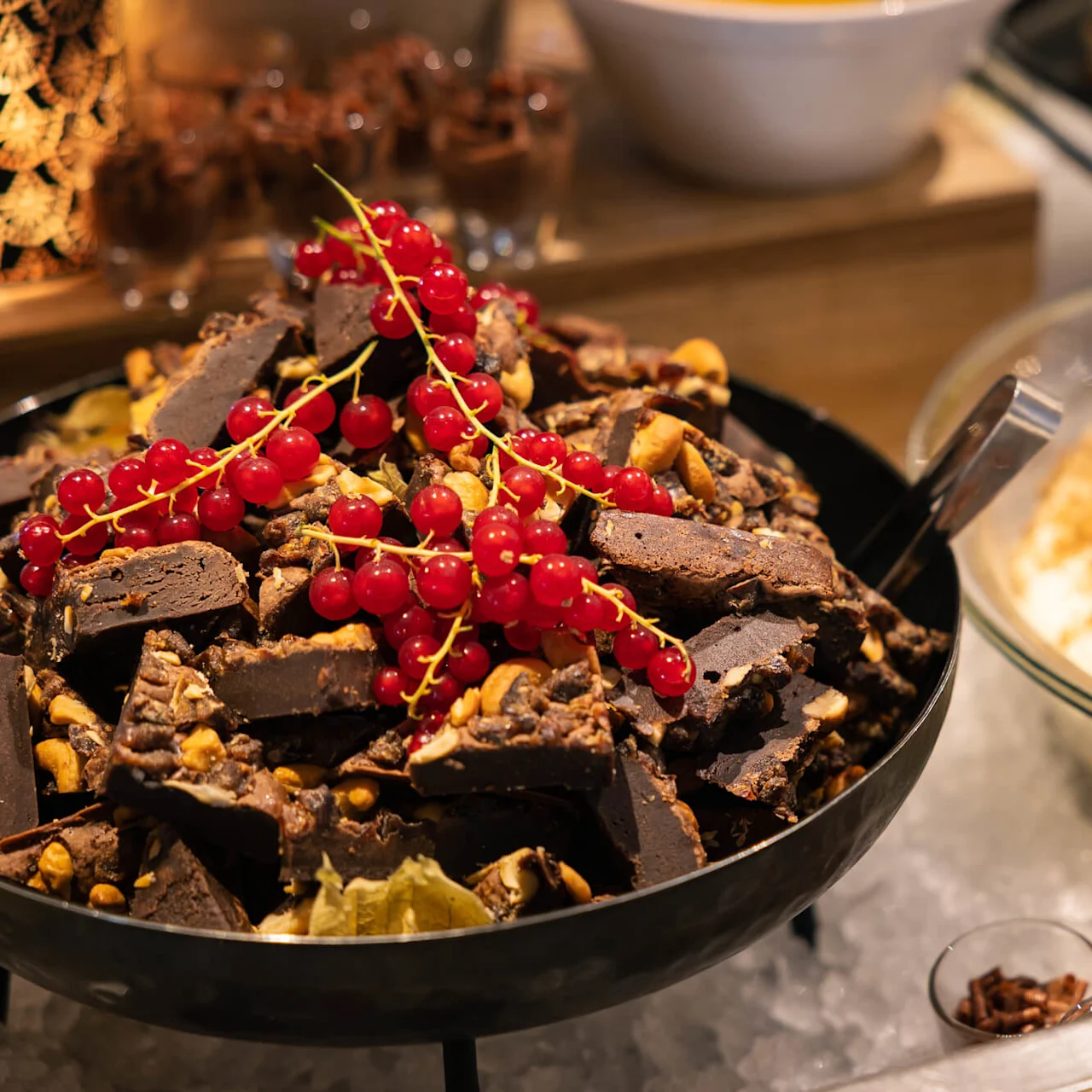 Sjokoladebiter toppet med rips i en bolle; i nærheten er det en glasskål med en dessert med jordbær og småkaker. Innstillingen ser ut til å være en buffet eller dessertutstilling.