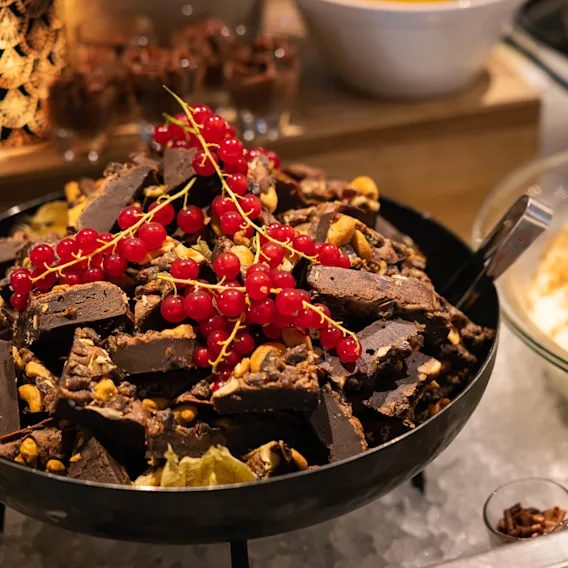 Sjokoladebiter toppet med rips i en bolle; i nærheten er det en glasskål med en dessert med jordbær og småkaker. Innstillingen ser ut til å være en buffet eller dessertutstilling.