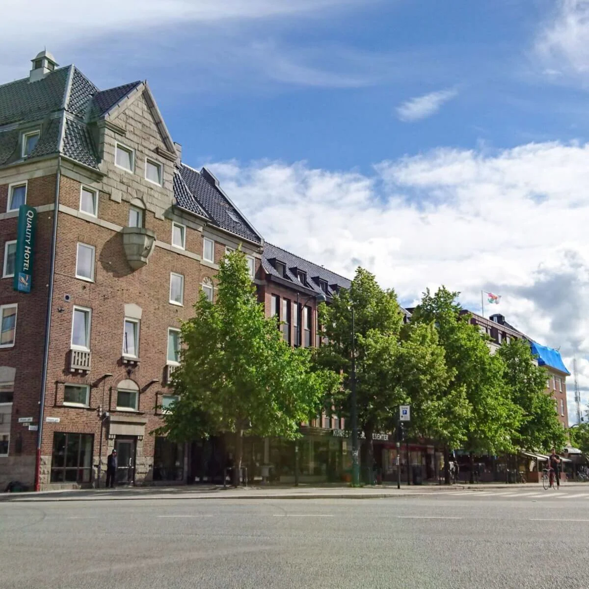 Et murstenshotel med tegltag står på et gadehjørne. En blå himmel med skyer og træer langs vejen. Skilte med teksten "Quality Hotel" og "Kurs & konferansesenter."