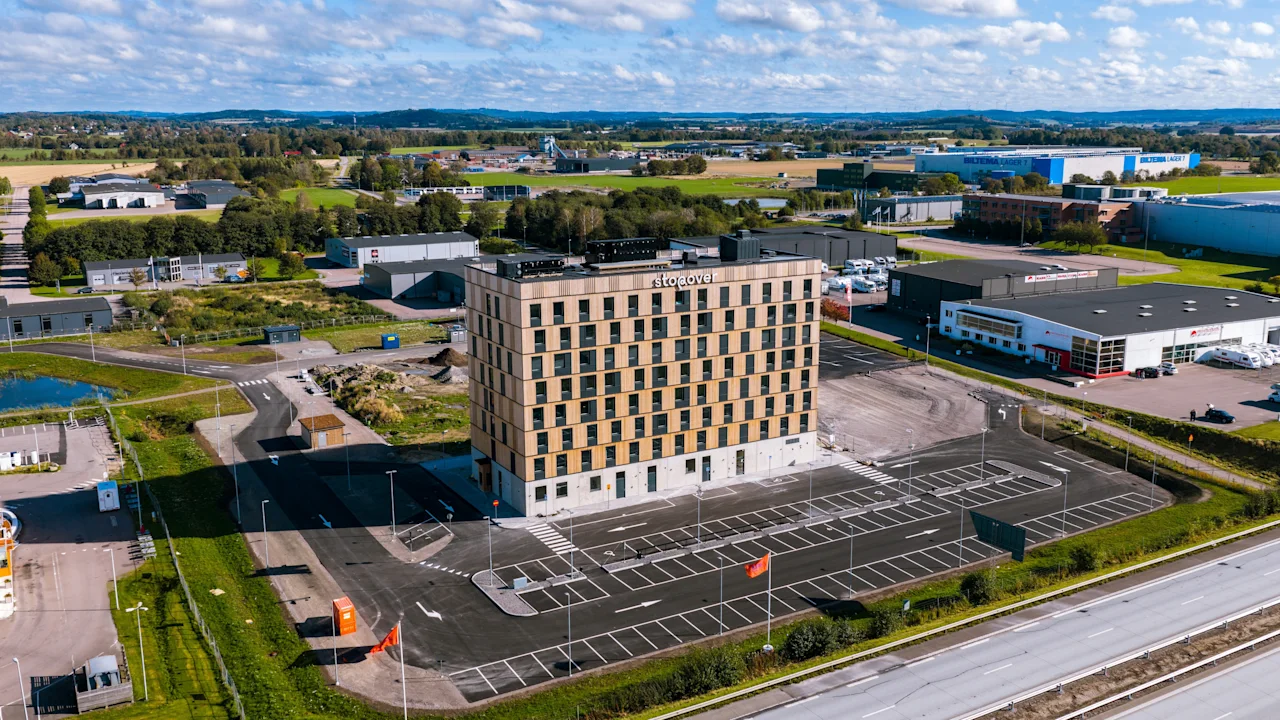 Ett högt, rektangulärt hotell med namnet "The Urban House" står på en stor parkeringsplats, omgiven av ett industriellt landskap med spridda lager och gröna fält under en delvis molnig himmel.