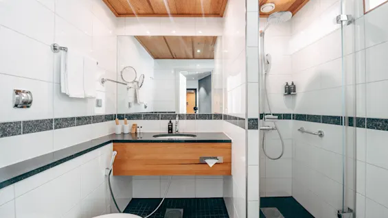 A modern bathroom with a wooden vanity holding a sink, mirrors above. To the right, a glass-enclosed shower with white walls and black tile floor. Ceiling has spotlights.