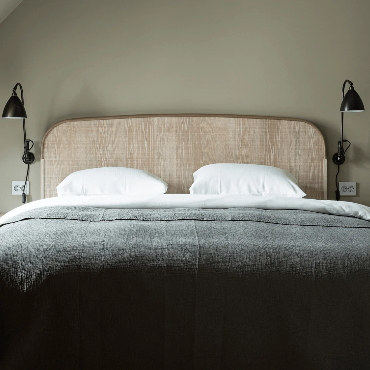 A bed with a wooden headboard and gray bedding, flanked by two black wall-mounted lamps, is centered in a small, minimalist room.