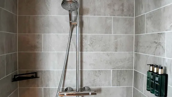 A modern shower with a rainfall and handheld head; set against gray tiled walls. Shampoo bottles are on a wall shelf. 