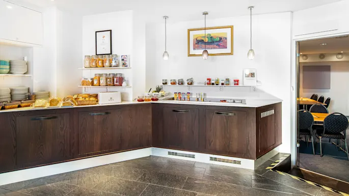Breakfast buffet with jars of cereals, bread, and condiments on a wooden counter. Plates stack to the left. Three pendant lights hang above. A framed picture decorates the wall.