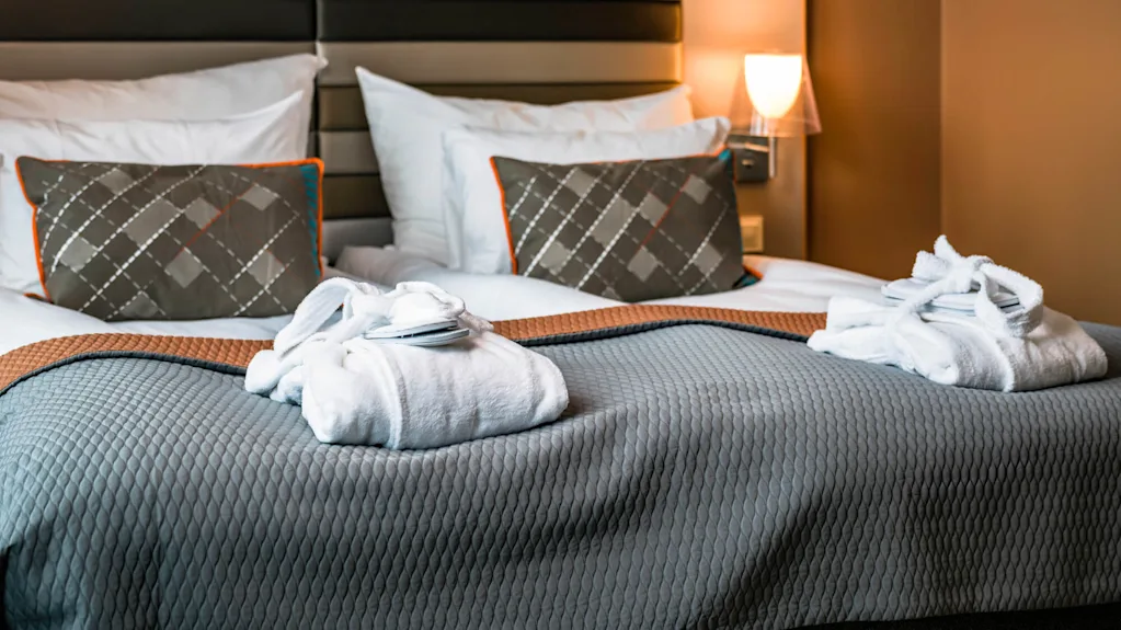 To hvide badekåber ligger pænt på en seng med et gråt tæppe i et varmt oplyst hotelværelse med mønstrede puder og en polstret sengegavl, der skaber en hyggelig atmosfære.