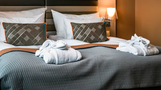 Two white bathrobes lie neatly on a bed with a gray quilt, in a warmly lit hotel room featuring patterned pillows and a padded headboard, creating a cozy atmosphere.