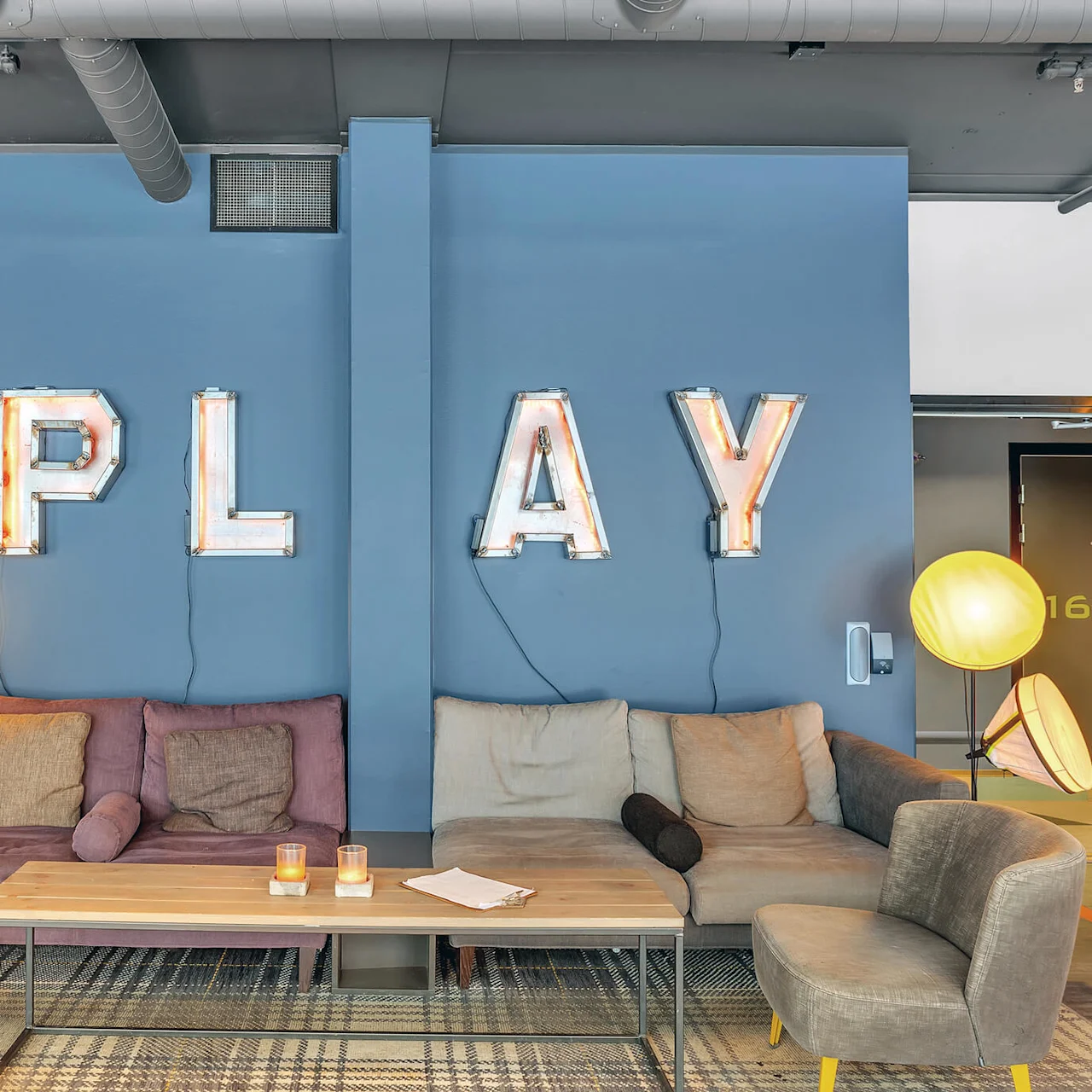 Couches sit against a blue wall with large illuminated letters spelling "PLAY". Warm floor lamps and a small coffee table create a cozy living space.