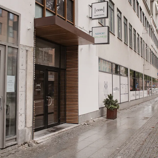 Ingång med glasdörrar märkta "Home Hotel" och "Odin's Hotel & Spa" vid en stadstrottoar, omgiven av ett träd och en rad fönster.