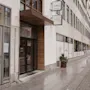 Ingång med glasdörrar märkta "Home Hotel" och "Odin's Hotel & Spa" vid en stadstrottoar, omgiven av ett träd och en rad fönster.