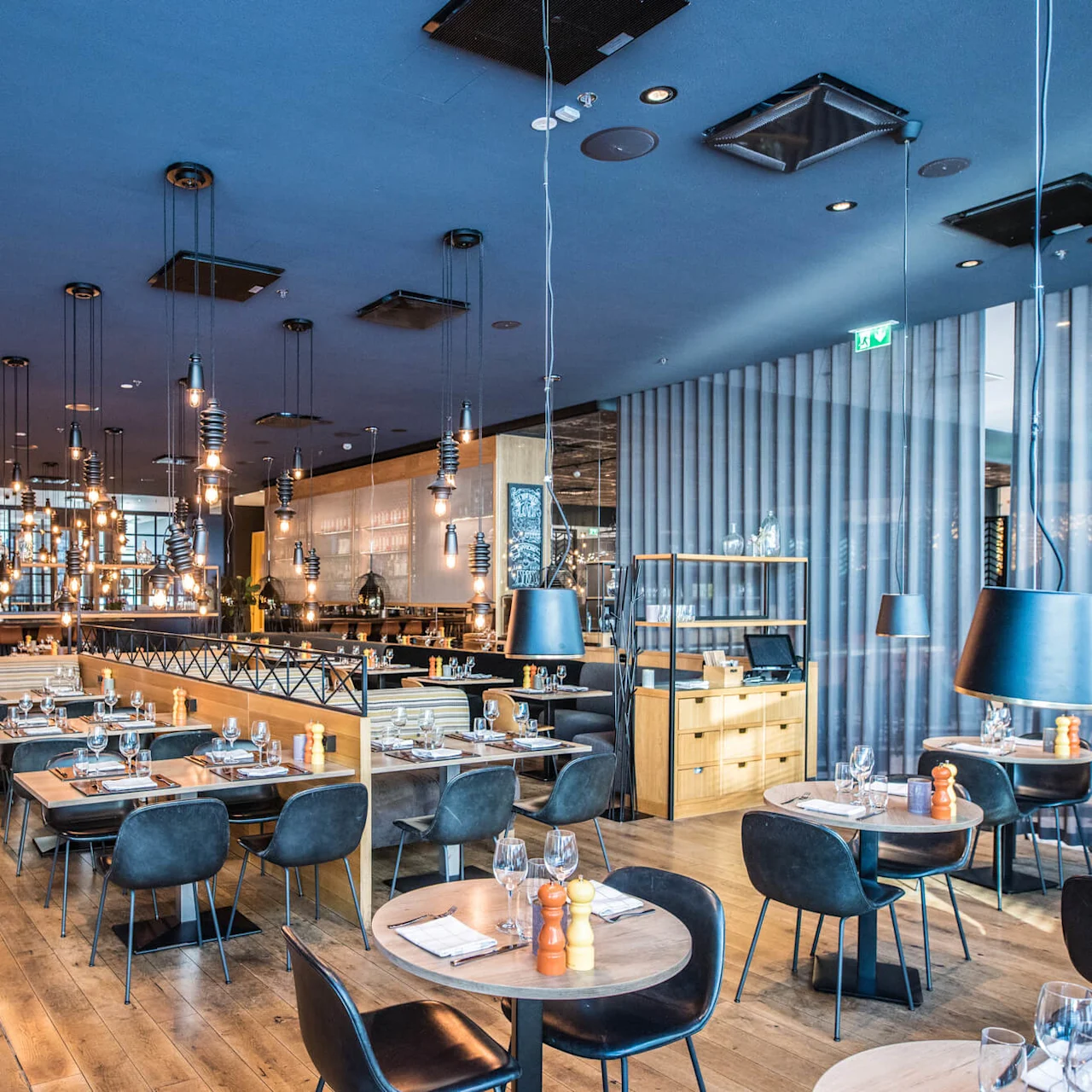 Elegant restaurant featuring neatly set tables with glasses and napkins. Modern lighting fixtures hang from the ceiling, illuminating the polished wooden floors. Shelves with decorative items line the back wall.