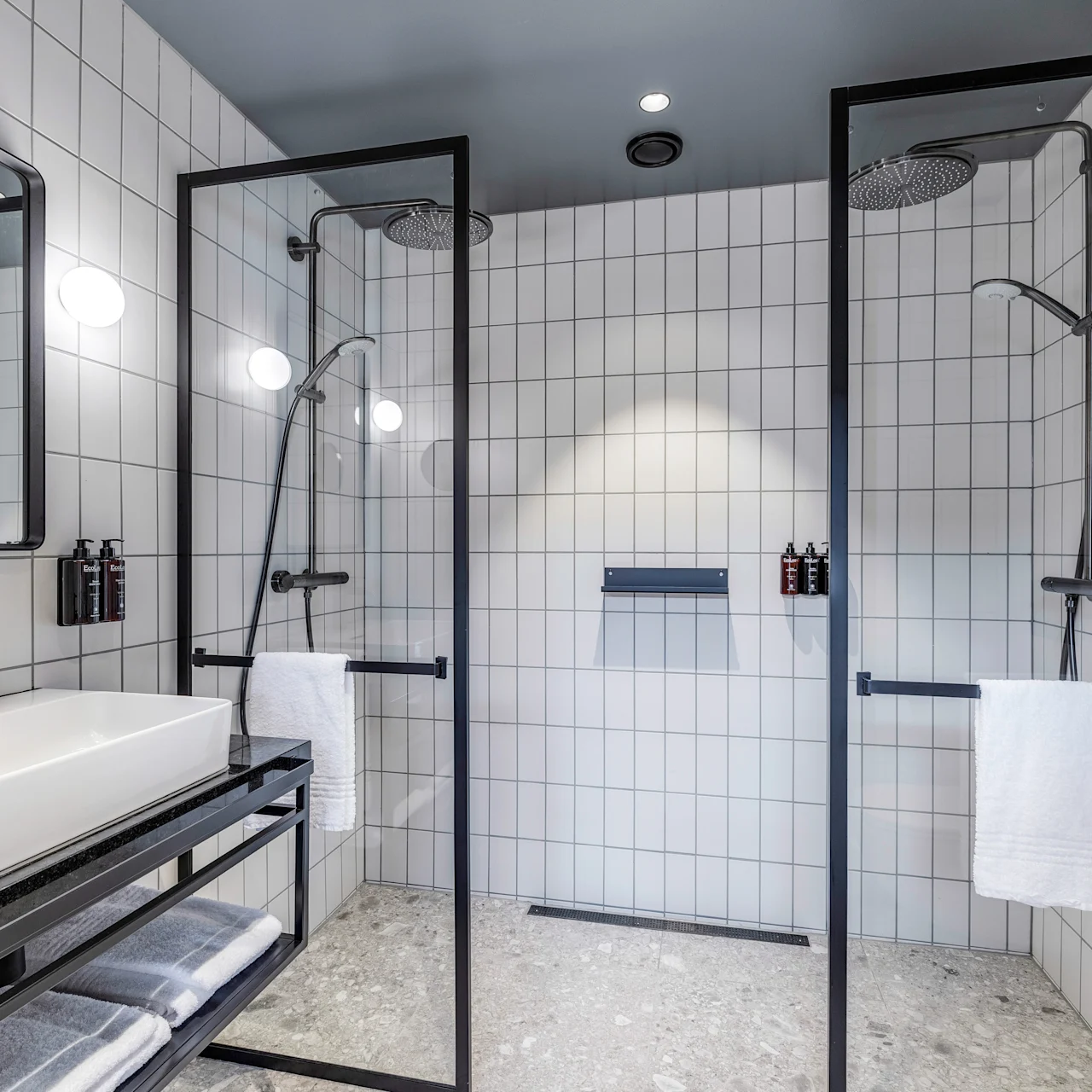 Hvidflisebelagt badeværelse med to brusekabiner og regnbrusere. Et rektangulært vask med metalhane står på en sort bordplade. Håndklæder og toiletartikler er pænt arrangeret.