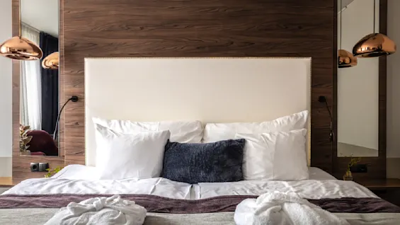 Ett mysigt hotellrum med en bekväm säng fylld med flera kuddar och utlagda morgonrockar, upplyst av stilrena överhängande lampor. De träfärgade detaljerna ger värme och elegans till rummet.