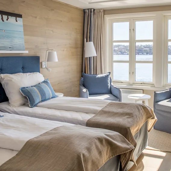 Et lyst hotellrom med to senger, lenestoler ved et stort vindu med utsikt over vann, og et skrivebordsområde.
