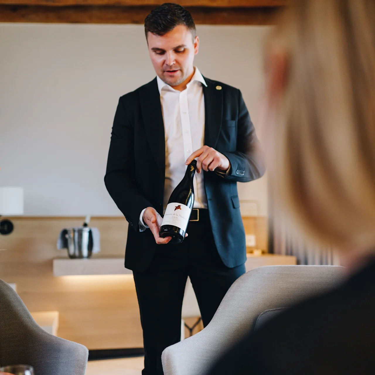 A man in a suit presents a wine bottle to seated guests in a warmly-lit room with wooden beams and modern furnishings.