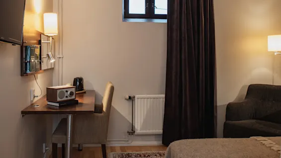 A comfortable hotel room featuring a desk with a radio and books, a cozy bed, and a window with dark curtains. Home Hotel Kung Oscar.