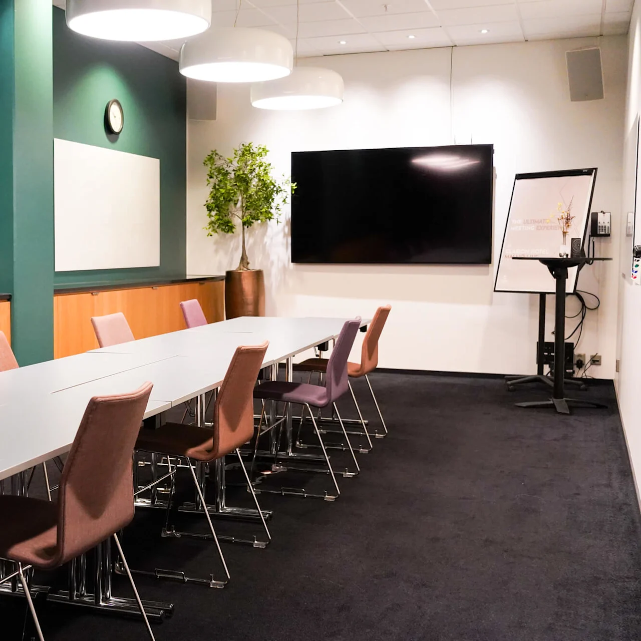 Konferansebord omgitt av stoler, plassert under tre taklamper i et møterom med en plante, stor skjerm og en flippover. Tekst: "THE ULTIMATE MEETING EXPERIENCE - HOTEL".